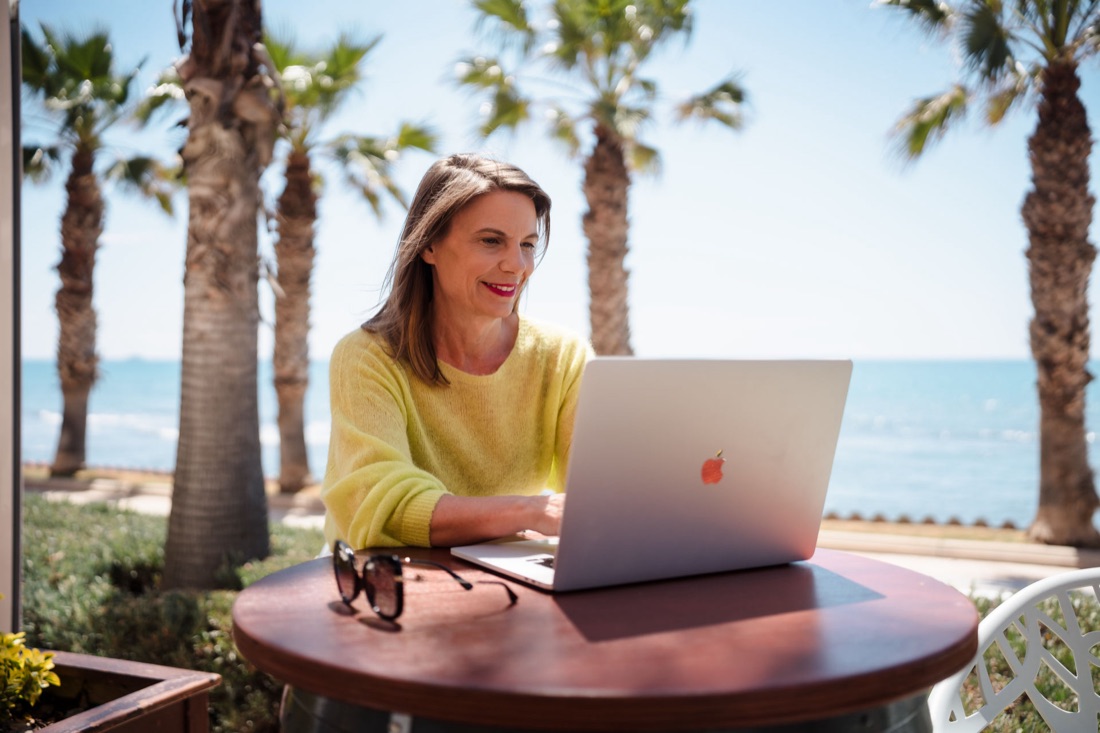 Sonja Kreye arbeitet lächelnd am Laptop unter Palmen